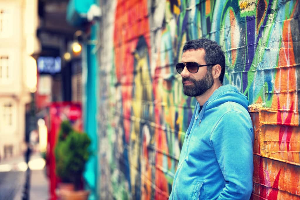 Bearded man in sunglasses and a blue hoodie leaning against a colorful graffiti wall on a city street.