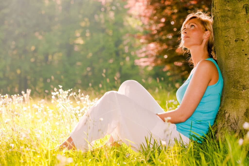 Woman in blue tank top relaxing against a tree in a sunlit meadow of wildflowers.