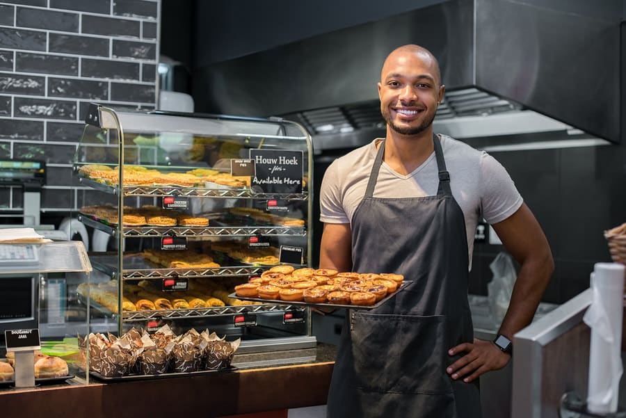 Smiling baker in apron holds tray of fresh pastries beside a bakery display case.