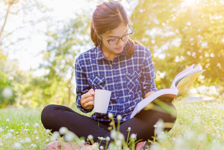 Woman sitting on green grass holding a cup of coffee while reading a book.