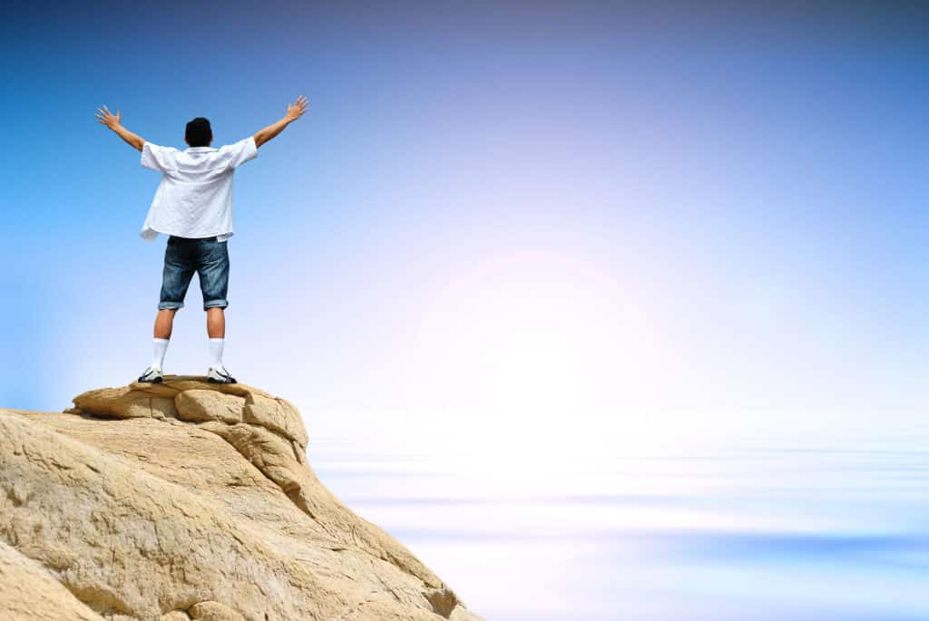 Man standing triumphantly with arms raised on a mountain summit at sunrise.