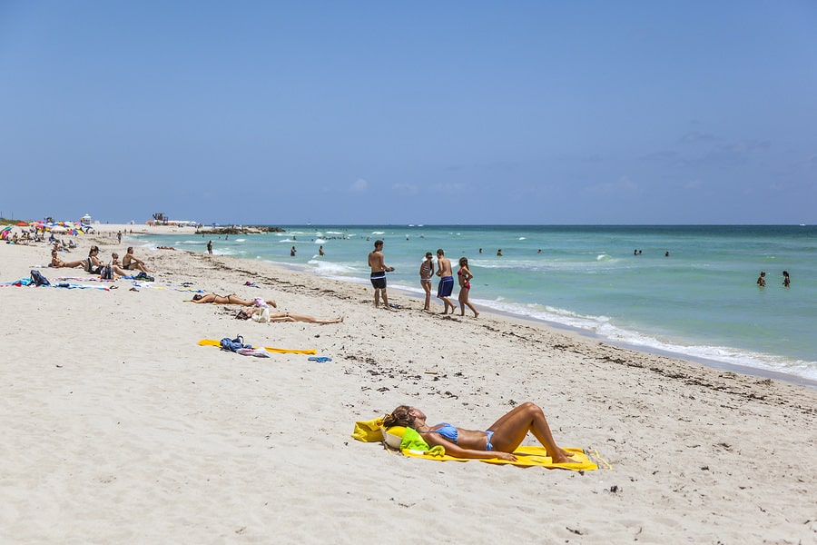 Sunbathers and swimmers enjoy a sunny sandy beach with clear blue ocean water under a bright sky.