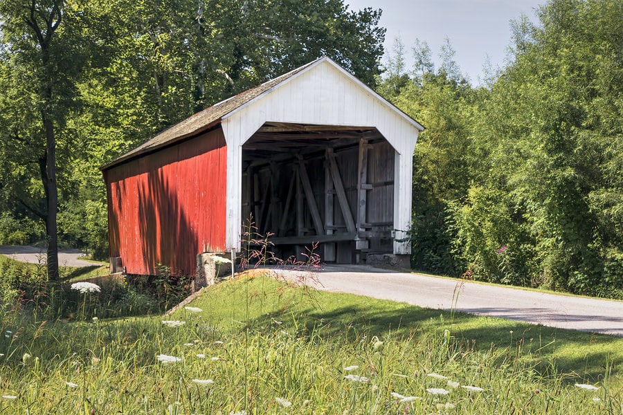 Red and white covered wooden bridge over a rural road surrounded by green trees and wildflowers.