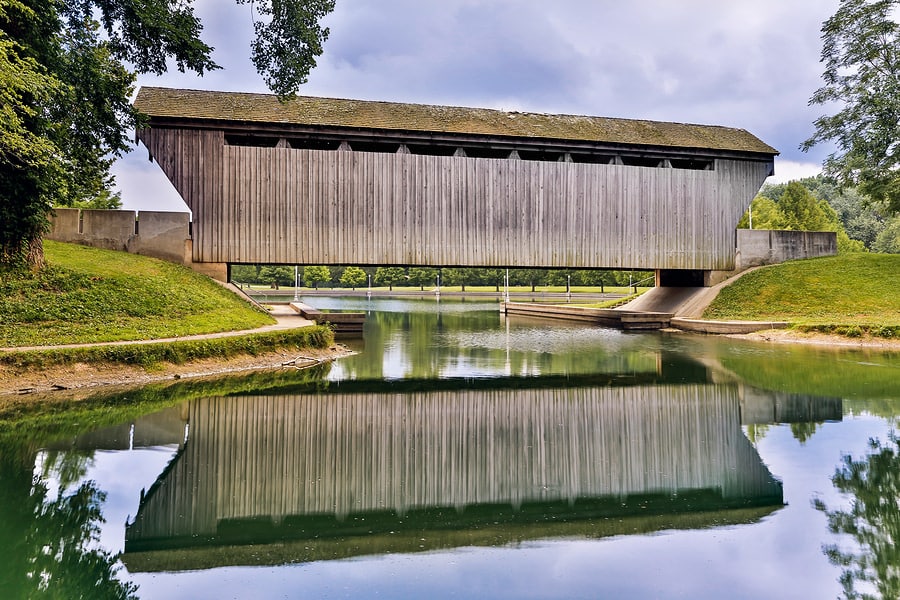 Wooden covered bridge reflected in calm water, surrounded by green hillsides and trees under a cloudy sky.
